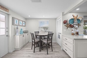 A kitchen with a dining table, chairs, cabinets, a window with blinds, and a decorative wall piece. - Casa Blanca - Villa B01 (Sarasota)