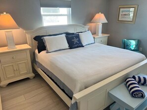 A neatly made bed with a white headboard, two bedside tables with lamps, a window with blinds, and a framed picture on the wall. - Casa Blanca - Villa B02 (Sarasota)