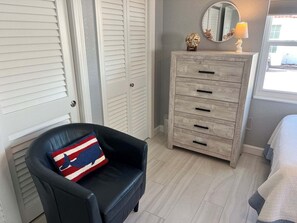 Ein Zimmer mit einem schwarzen Ledersessel, einem Holzschrank mit Schubladen, einem runden Spiegel und einer Lampe auf dem Schrank.
