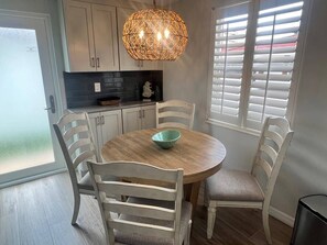 A dining area with a round table, white chairs, a hanging light fixture, and a window with blinds. - Casa Blanca - Villa B04 (Sarasota)