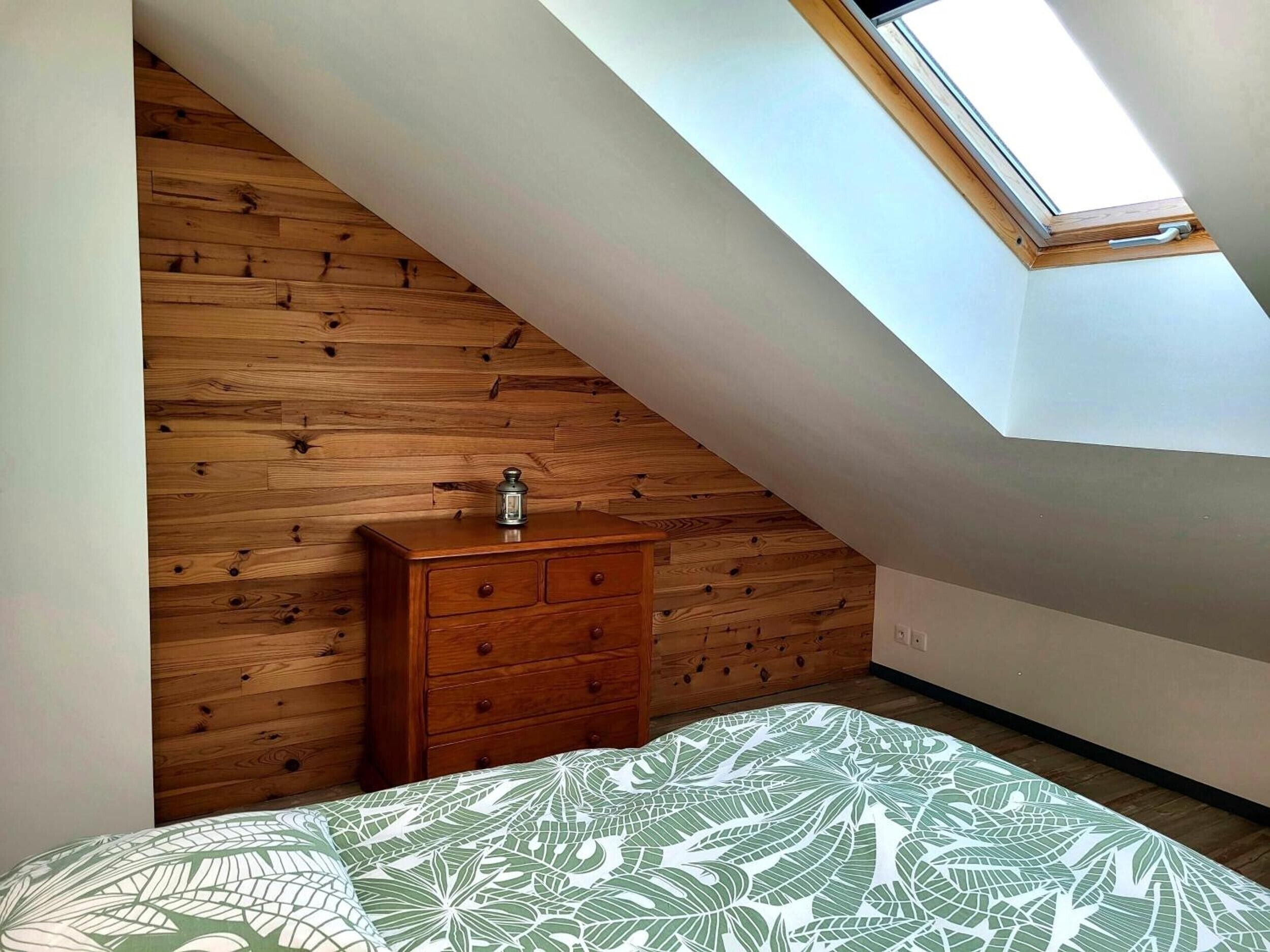 Une chambre avec un plafond en pente, un mur lambrissé, une commode, un lit recouvert d’une couvre-lit à motifs de feuilles et un puits de lumière.