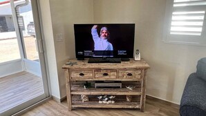 A television on a wooden entertainment center with a remote control, a small decorative item, and a window with blinds. - Casa Blanca - Villa B10 (Sarasota)