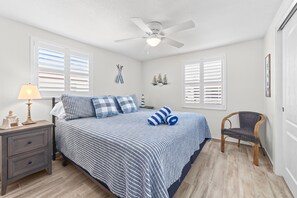 A bedroom with a bed, a nightstand, a lamp, a chair, a ceiling fan, and a window with blinds. - Casa Blanca - Villa B10 (Sarasota)
