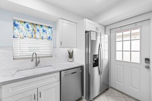A modern kitchen with white cabinetry, stainless steel appliances, and a window with a floral curtain. - Casa Blanca - Villa B11 (Sarasota)