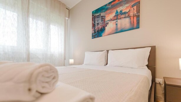 Un letto rifatto con lenzuola bianche, un quadro raffigurante una veduta cittadina sulla parete e una finestra con tende leggere.