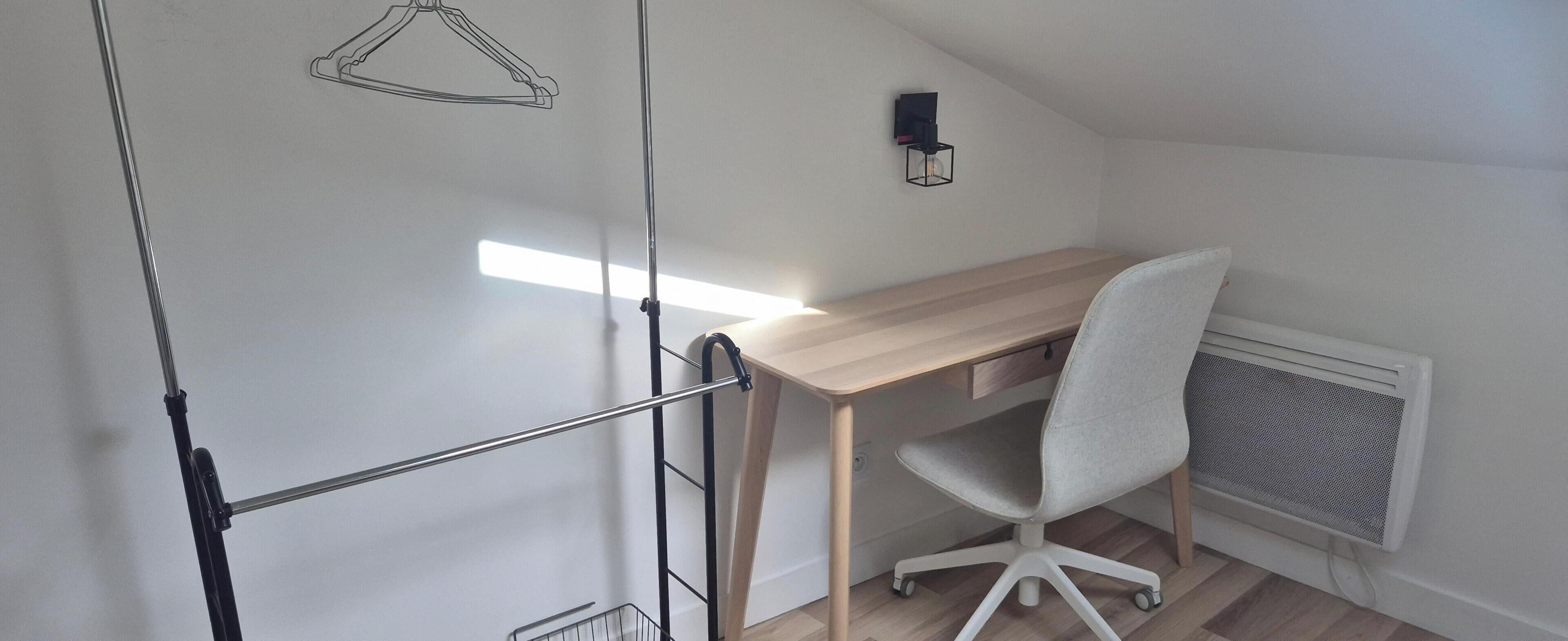 Ett rum med ett skrivbord i trä, en vit kontorsstol, ett element och en väggmonterad lampa.