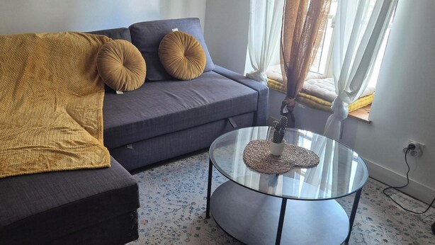 Un soggiorno con divano, tavolino in vetro, coperta e finestra con tende.