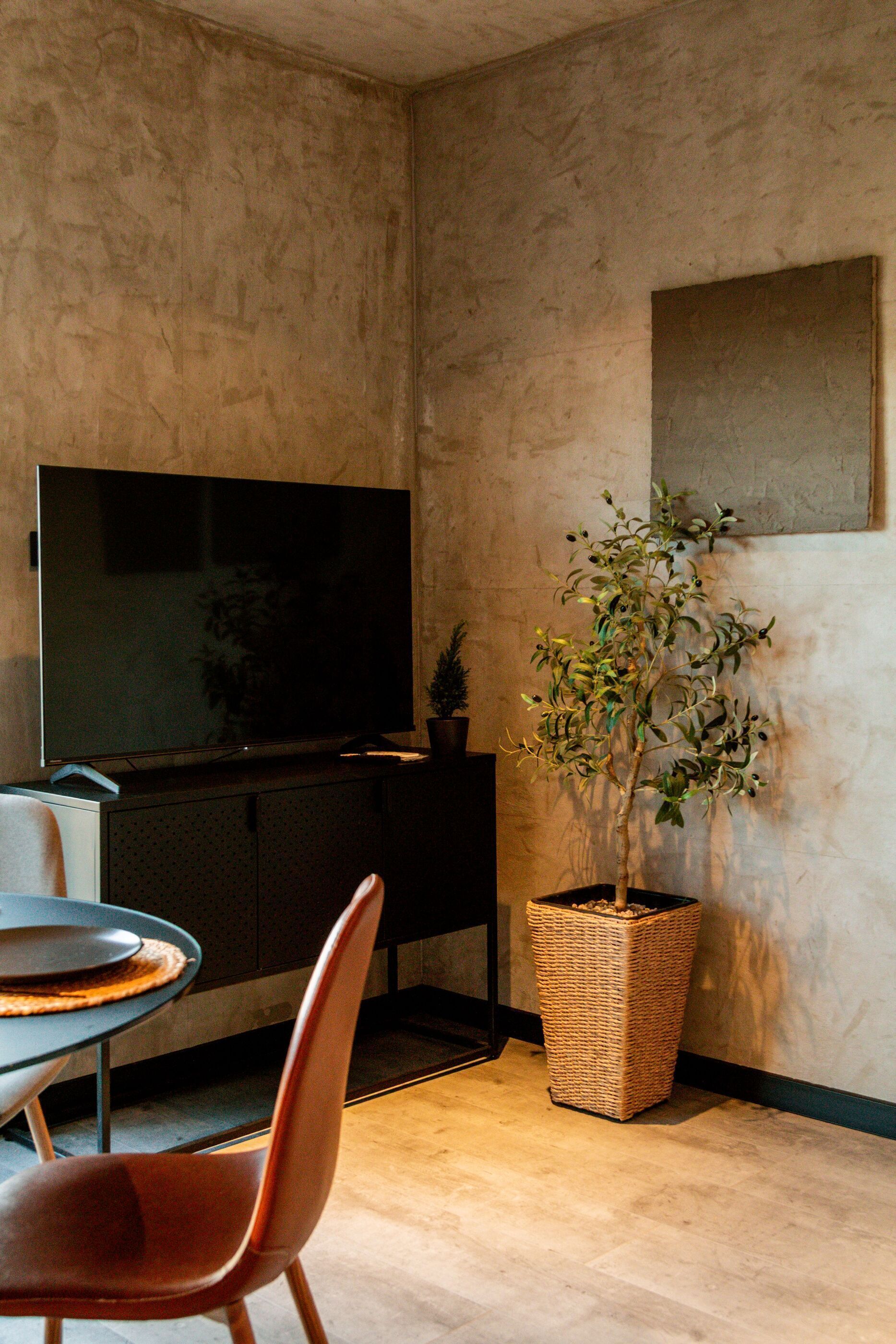 Ein modernes Wohnzimmer mit einem Flachbildfernseher, einem Esstisch mit Stühlen, einer Zimmerpflanze und einem großen Wandbild.