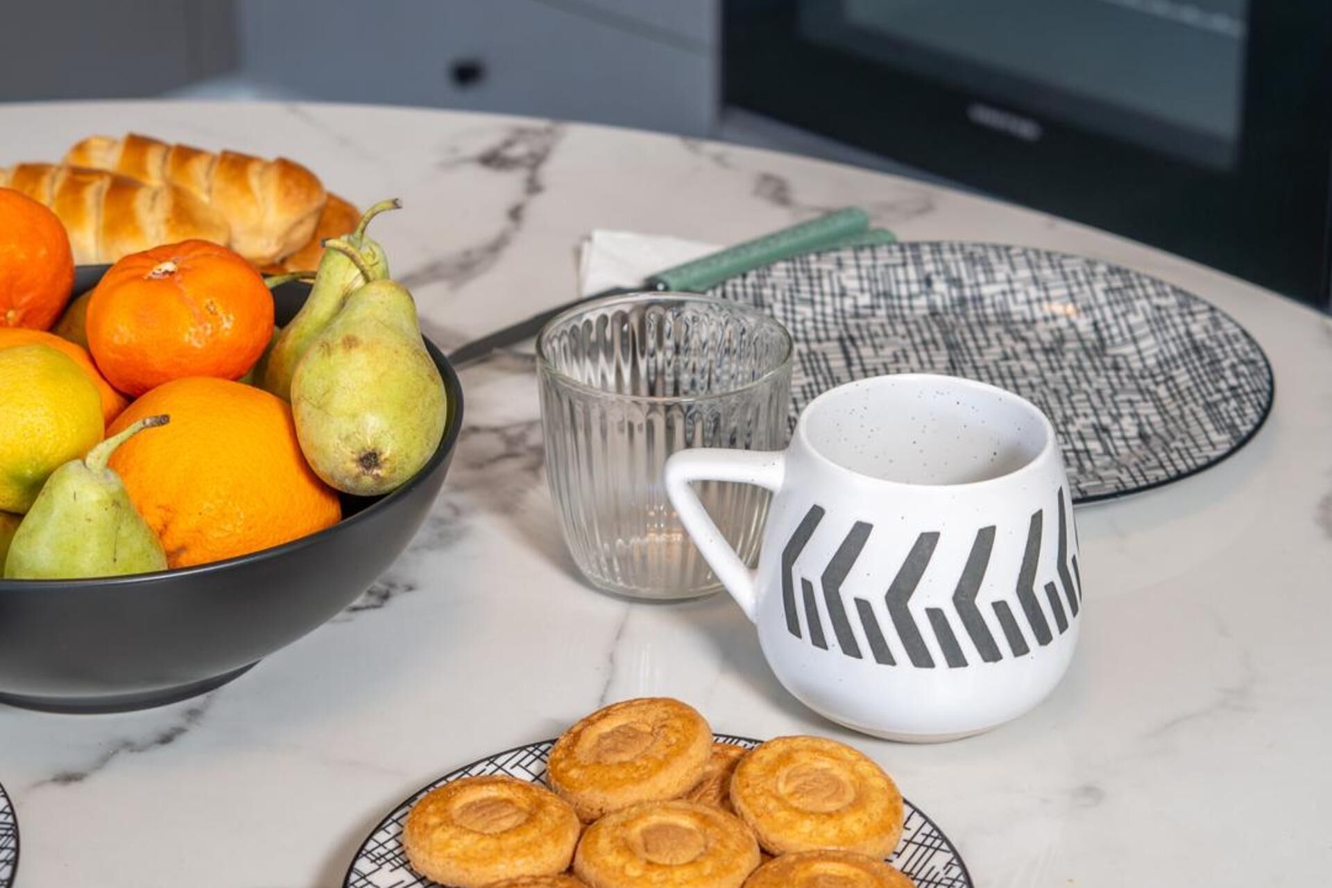 Mesa de café da manhã com uma tigela de frutas, um prato de doces, uma caneca de vidro e um jogo americano estampado.