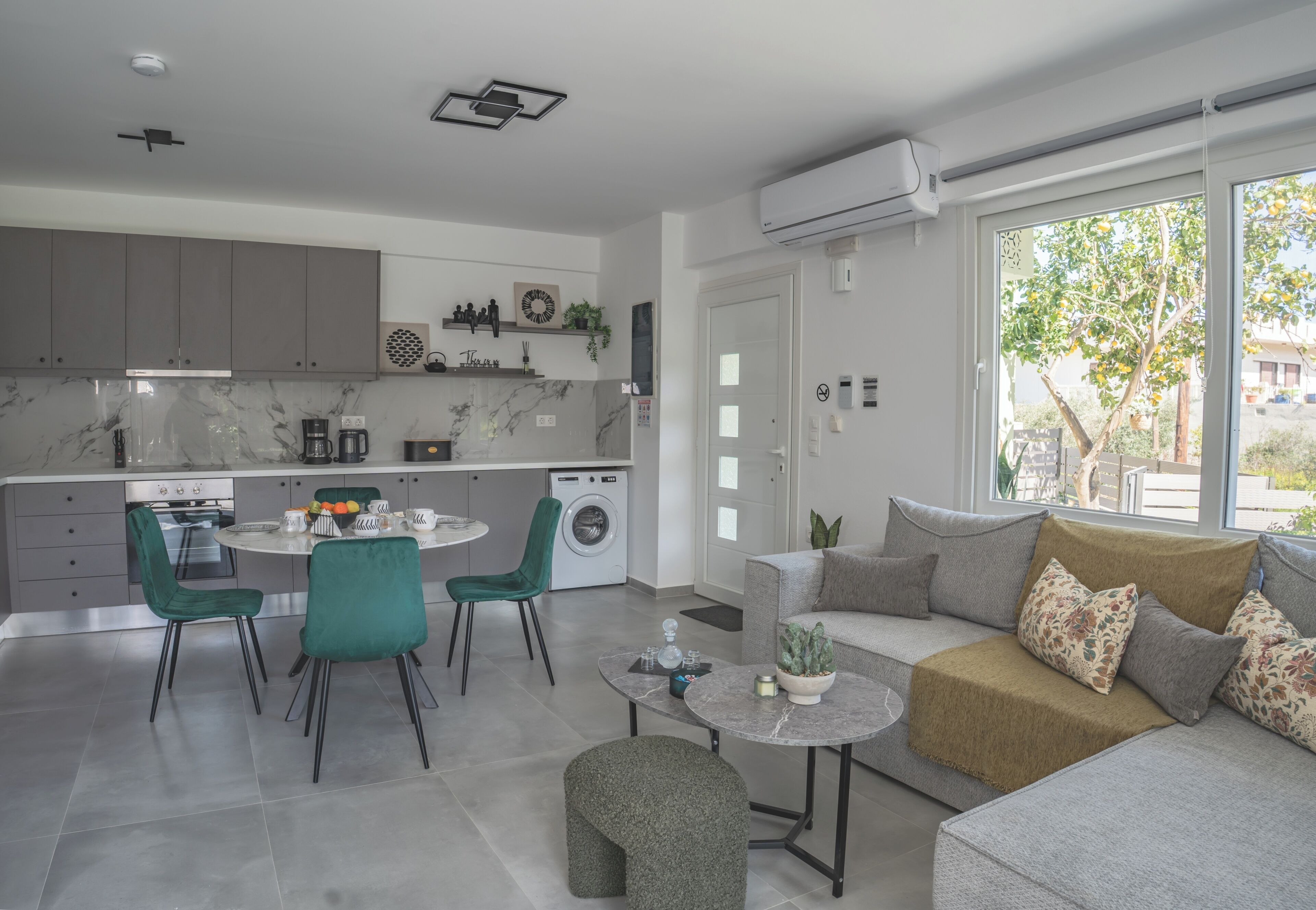 Cozinha moderna com armários cinza, área de refeições com mesa redonda e cadeiras, sofá com almofadas e uma janela grande com vista para área verde.