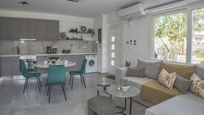 A modern kitchen with grey cabinets, a dining area with a round table and chairs, a sofa with cushions, and a large window with a view of greenery. - Thyme Aroma - Quiet Nature Patio (Rethymno)