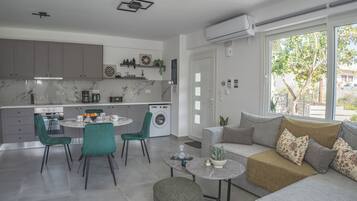 Cozinha moderna com armários cinza, área de refeições com mesa redonda e cadeiras, sofá com almofadas e uma janela grande com vista para área verde.