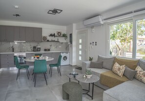 Eine moderne Küche mit grauen Schränken, ein Essbereich mit rundem Tisch und Stühlen, eine Couch mit Kissen und ein großes Fenster mit Blick auf Grünflächen.