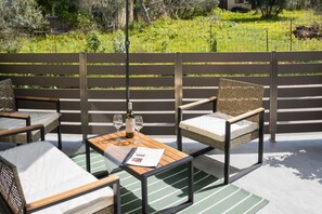 An outdoor seating area with wooden benches and woven chairs, a table with a bottle and glasses, and a backdrop of a garden with a fence. - Thyme Aroma - Quiet Nature Patio (Rethymno)
