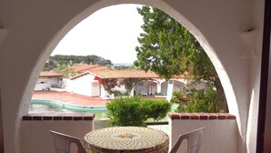 A balcony with a table and chairs, a view of a swimming pool, and a building with red-tiled roofs. - Family Oasis: Pool and Garden - Baja Sardinia (Arzachena)