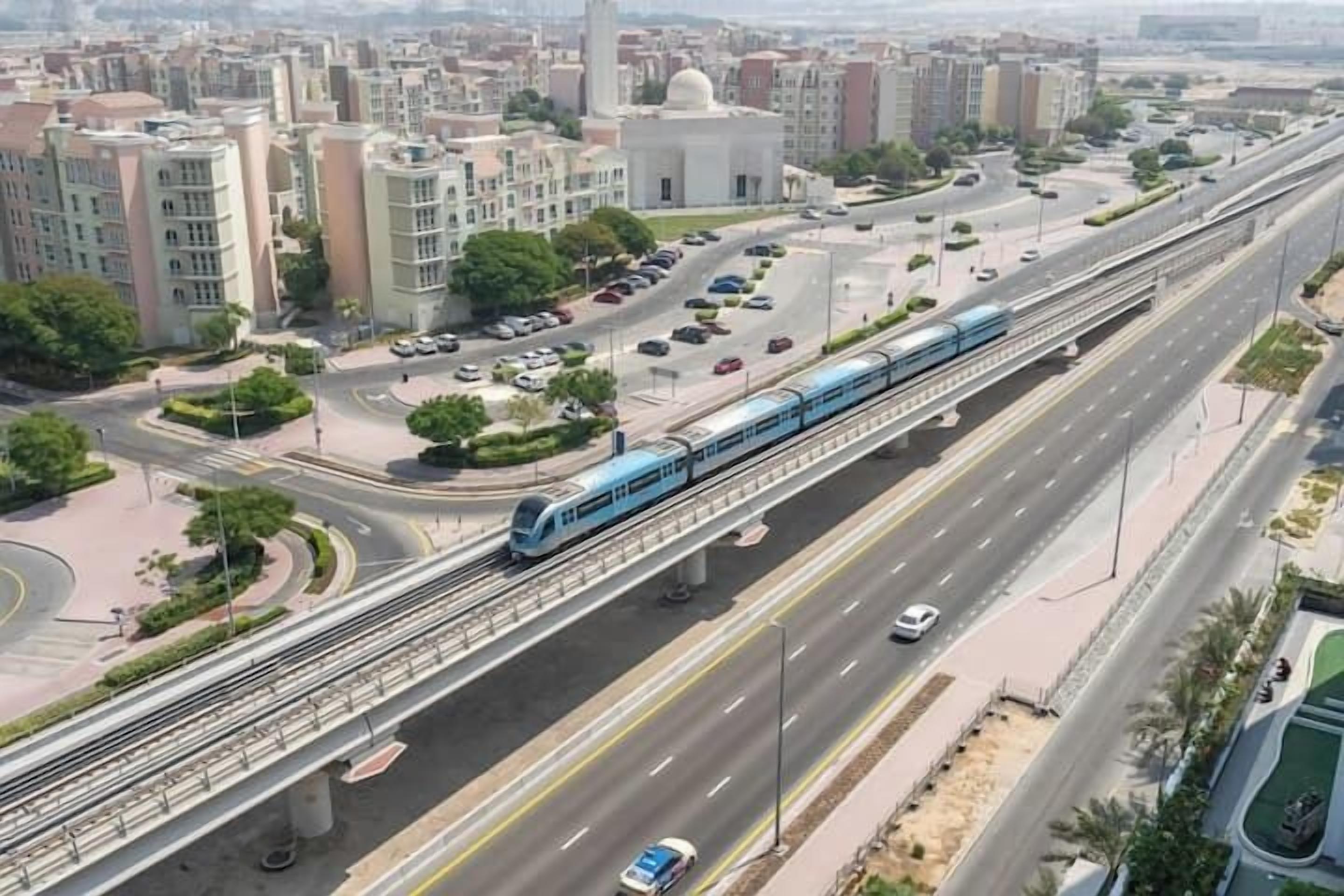 Un tren en una vía elevada, una carretera con varios carriles y edificios circundantes.