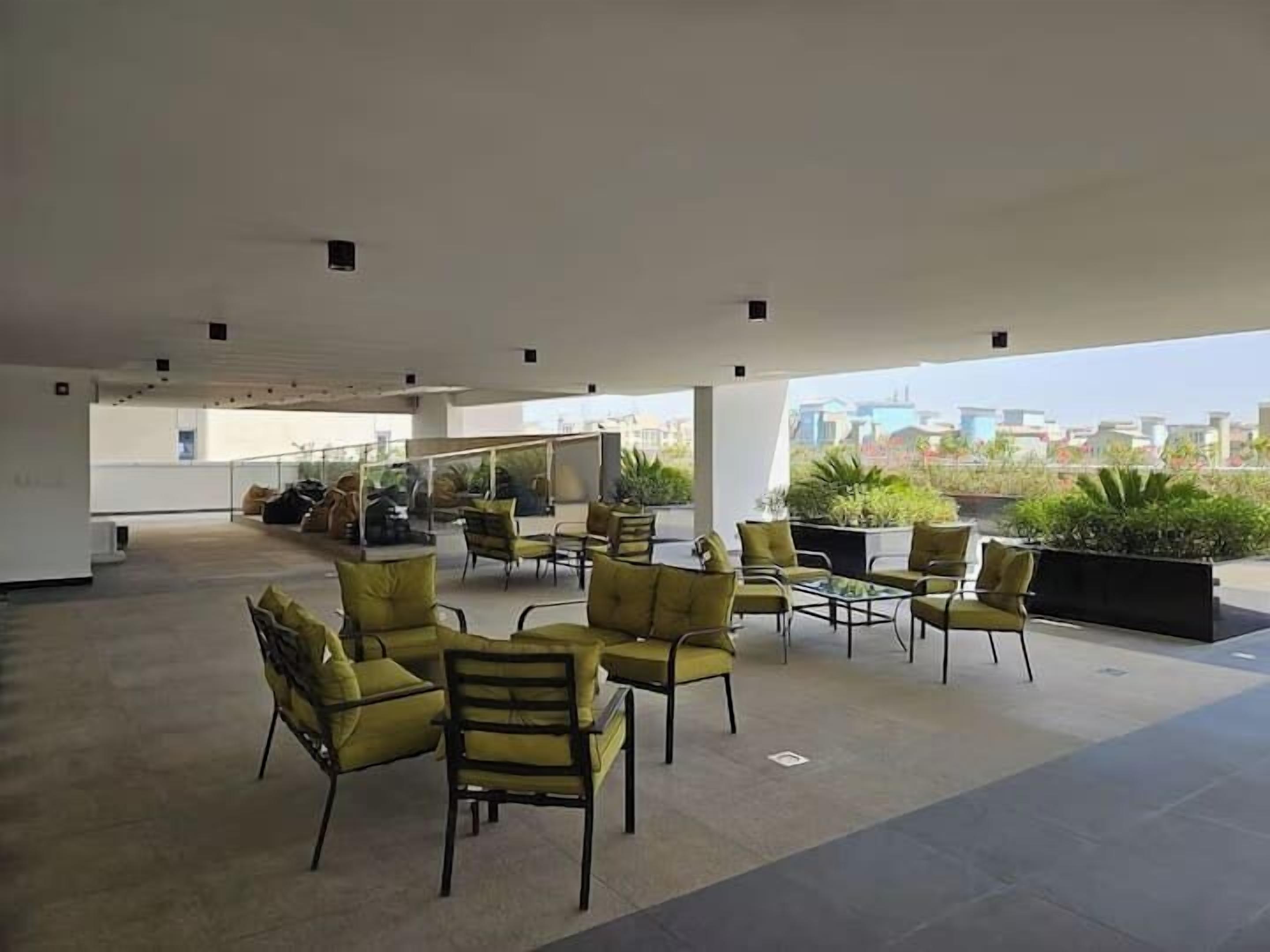 An outdoor seating area with yellow chairs and tables, surrounded by potted plants, with a view of buildings in the background.