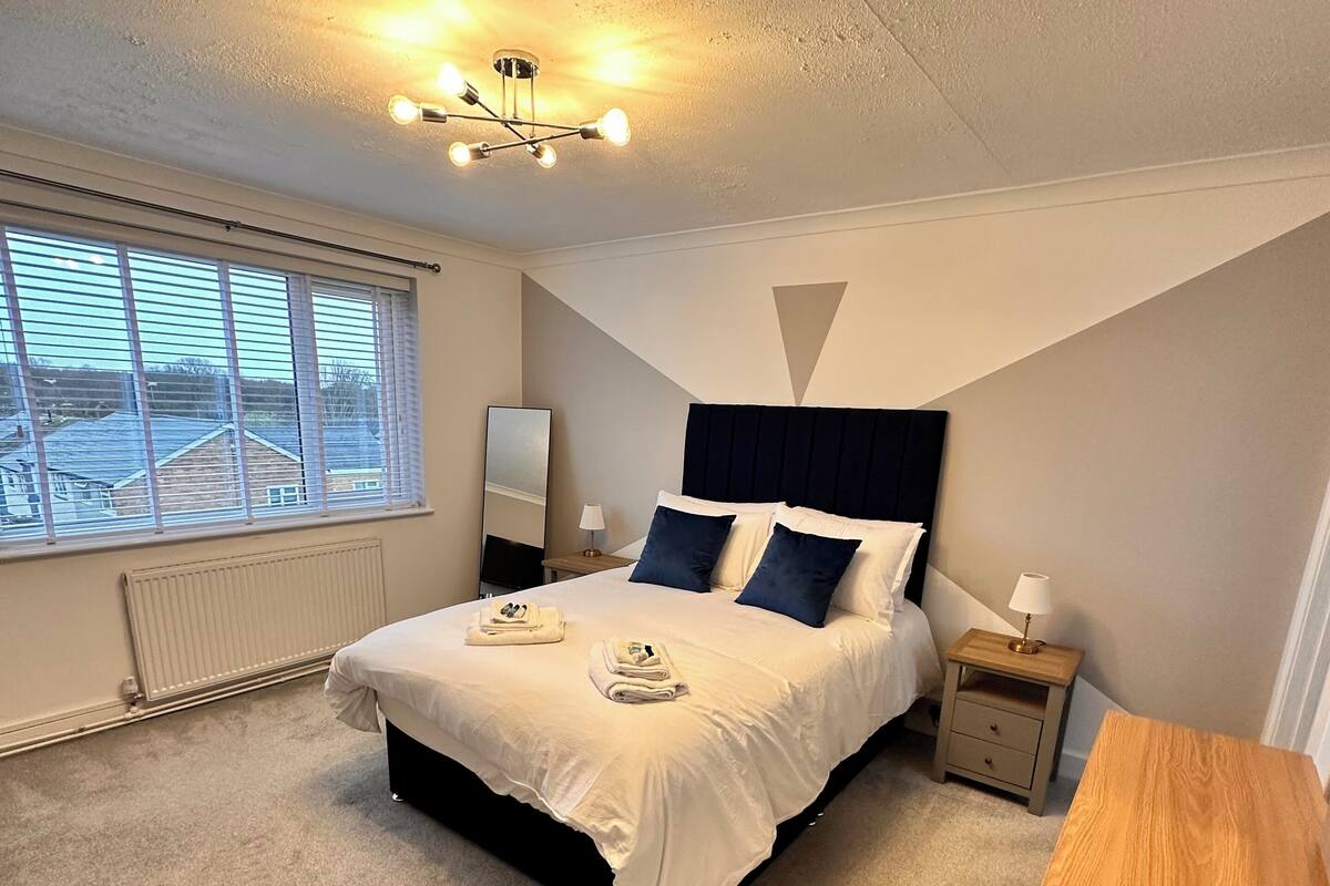 Une chambre à coucher comprenant un lit, des tables de chevet, un bureau, une fenêtre et un luminaire au plafond.