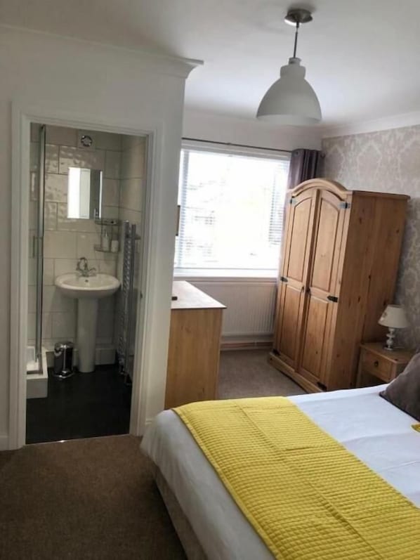 A bedroom with a bed, a wooden wardrobe, a bedside table with a lamp, a window with a view, and a bathroom with a sink and mirror. - The Poachers (Stevenage)