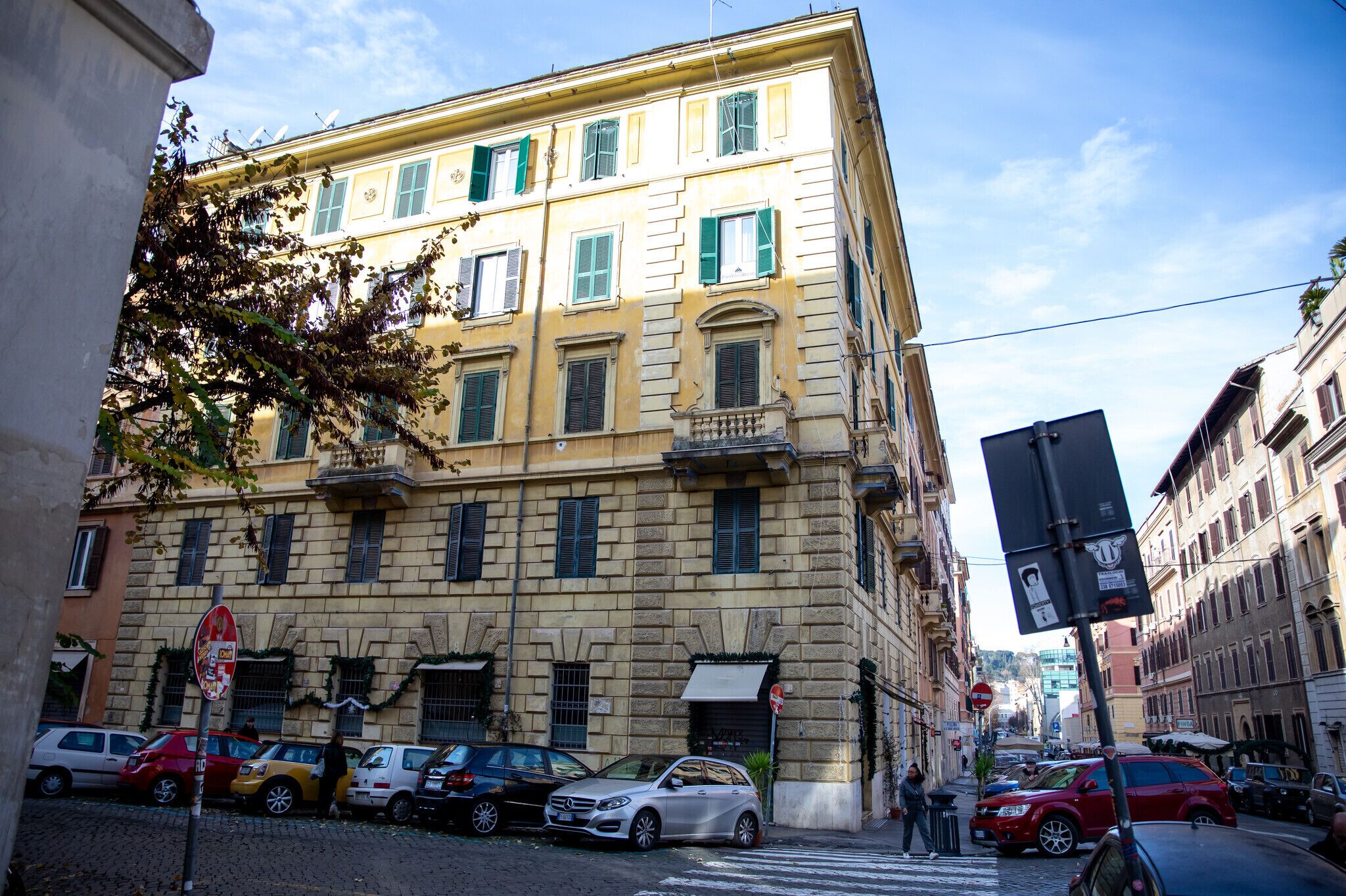 Ein mehrstöckiges Gebäude im klassischen Stil, mit einem Balkon im zweiten Stock, einem Eingang im Erdgeschoss und einer Straßenszene mit parkenden Autos und einer Fußgängerquerung.