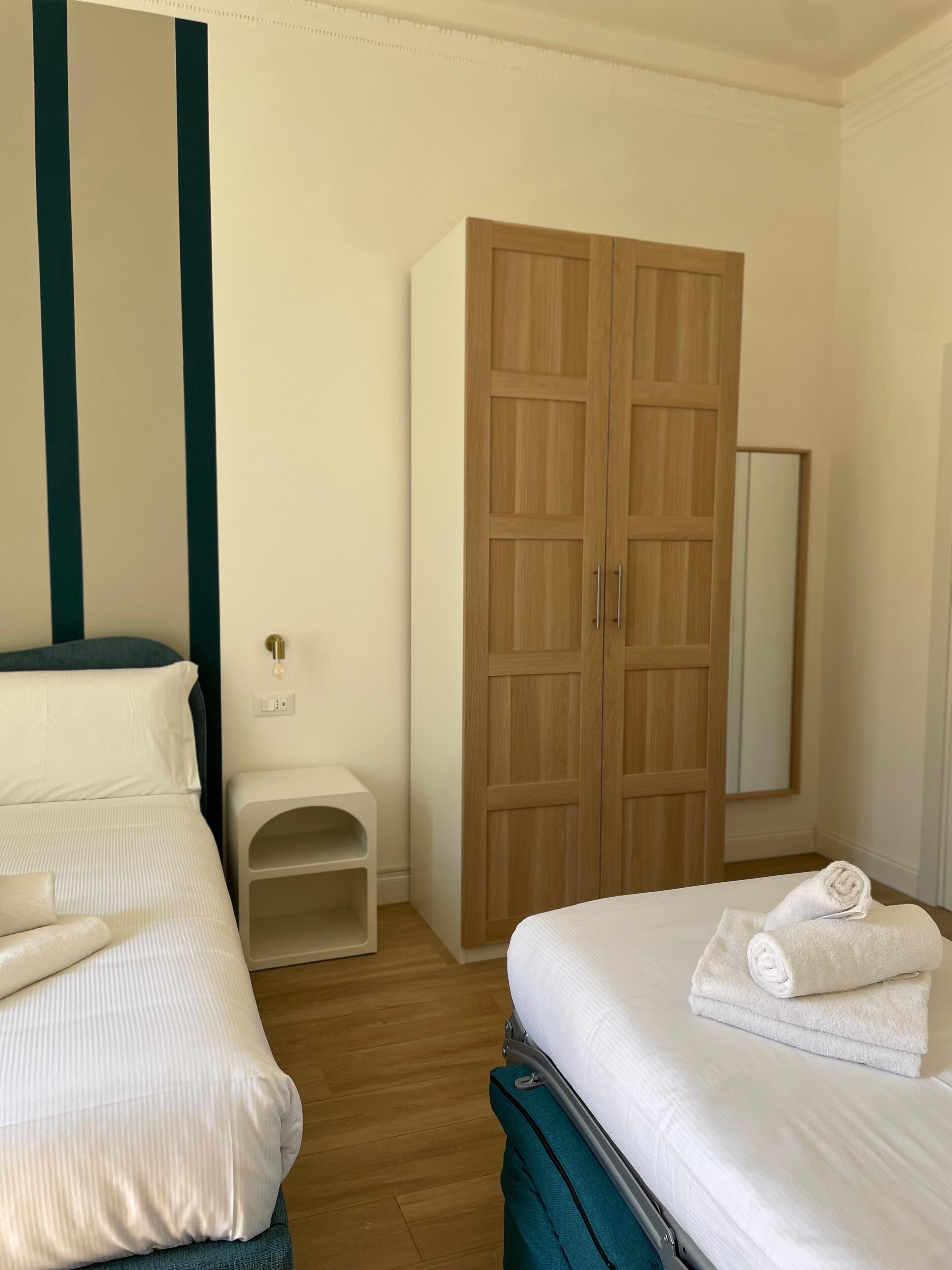 A hotel room with two beds, a wooden wardrobe, a small bedside table, and a mirror.