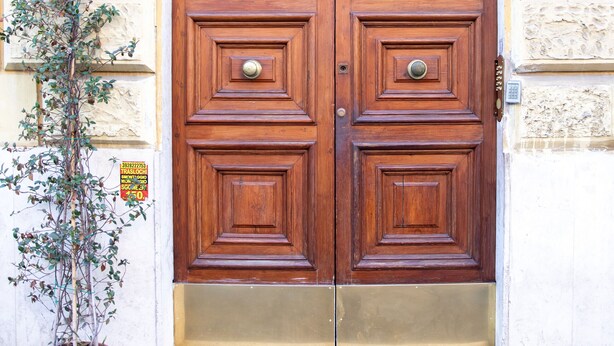 Une porte en bois à deux battants, ornée d’une plaque de frappe en laiton, un pot de fleurs placé à gauche, et un mur à texture, sur lequel est fixée une enseigne au-dessus de la porte.