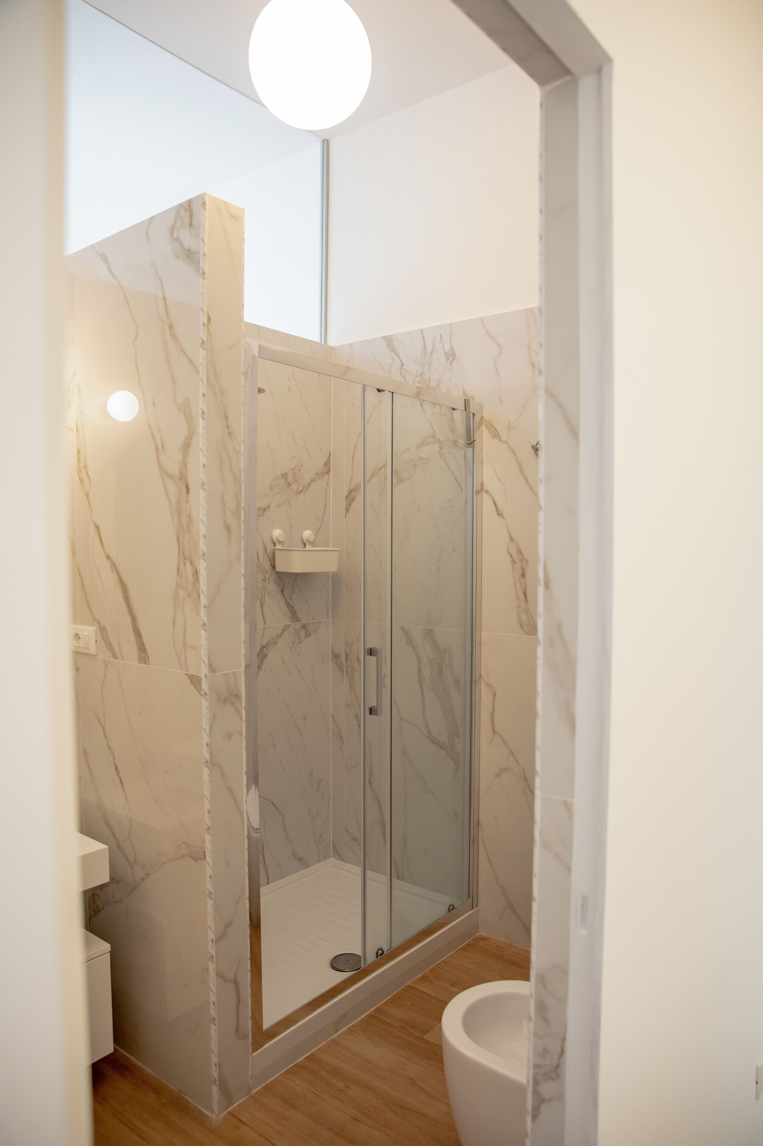 A modern bathroom with a glass-enclosed shower, marble walls, and a wooden floor.