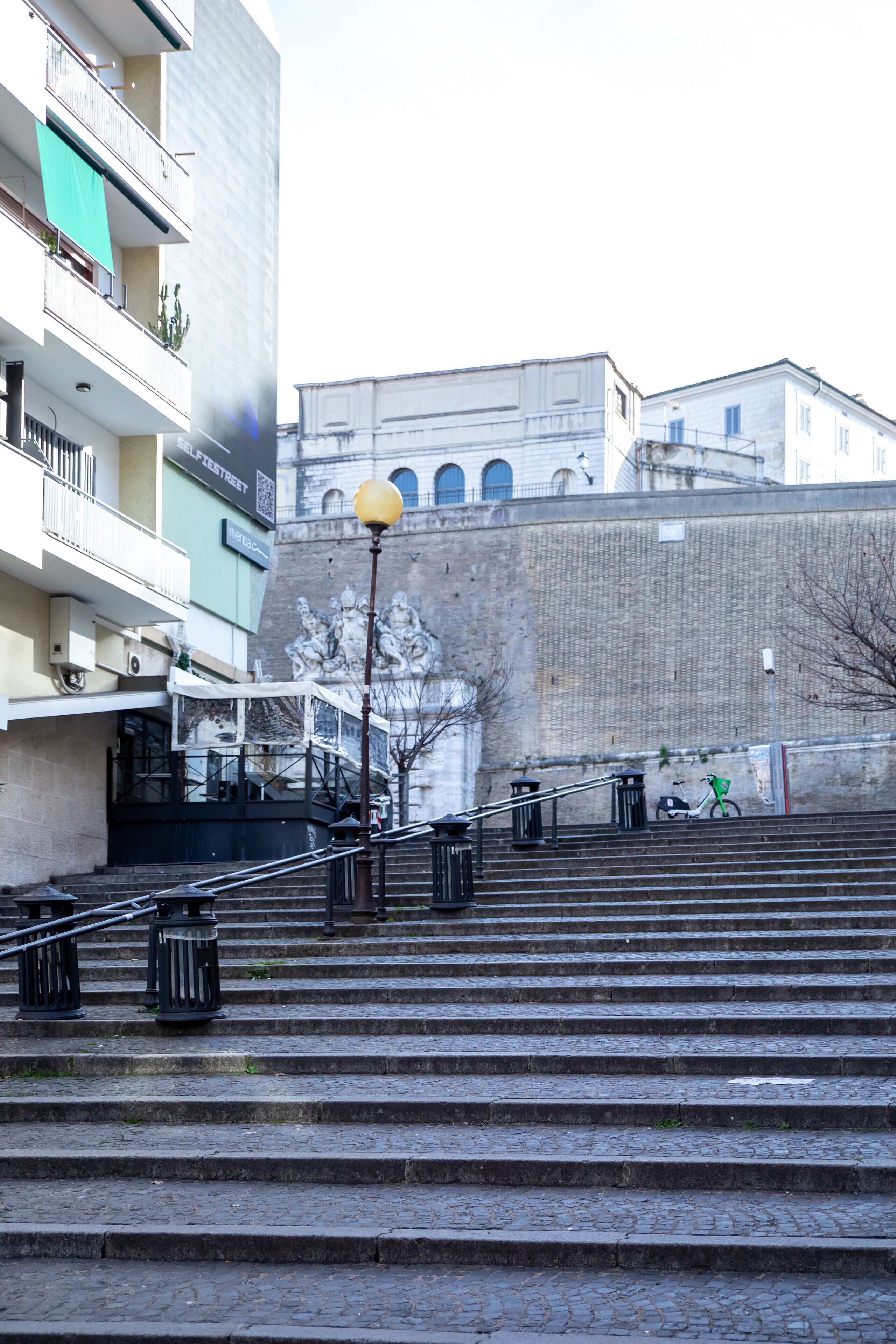 Un escalier en pierre mène à un bâtiment orné d’une fresque murale. Un lampadaire et un vélo sont également présents, ce dernier étant garé sur le côté.