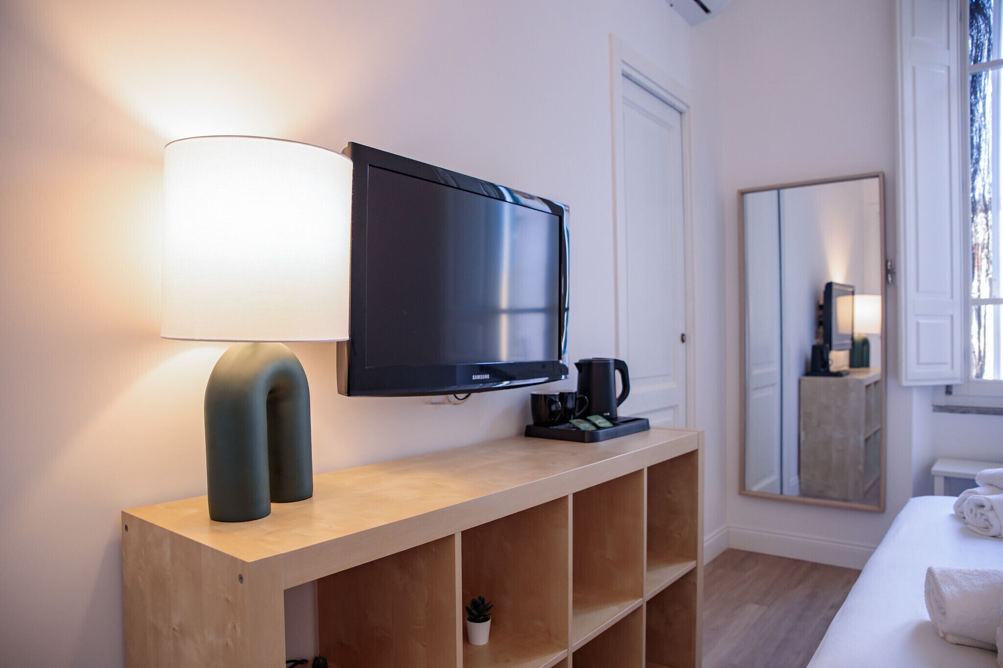 A modern hotel room with a flat-screen TV, a bedside lamp, a coffee maker, a mirror, and a neatly made bed.