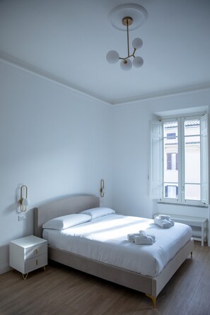 Een minimalistische slaapkamer met een bed, nachtkastjes, een radiator en een raam met open luiken.