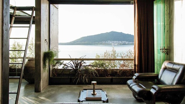A room with a view of a lake and a mountain, a green door, a black leather sofa, a small table with a book, and a ladder against the wall. - Luxury Waterfront Loft with Panoramic Views (Caminha)