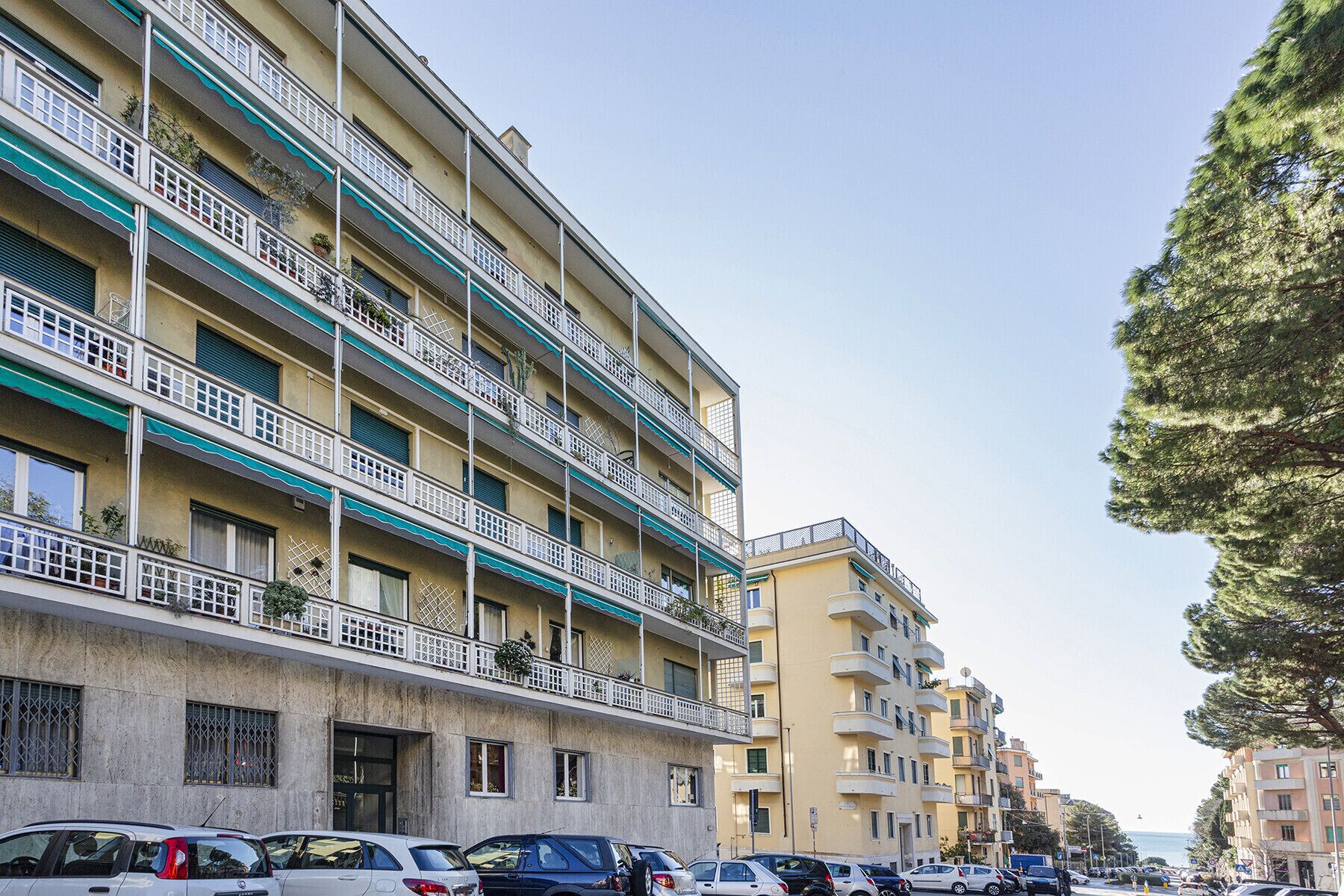A multi-story residential building with balconies, parked cars, and a tree on the right.