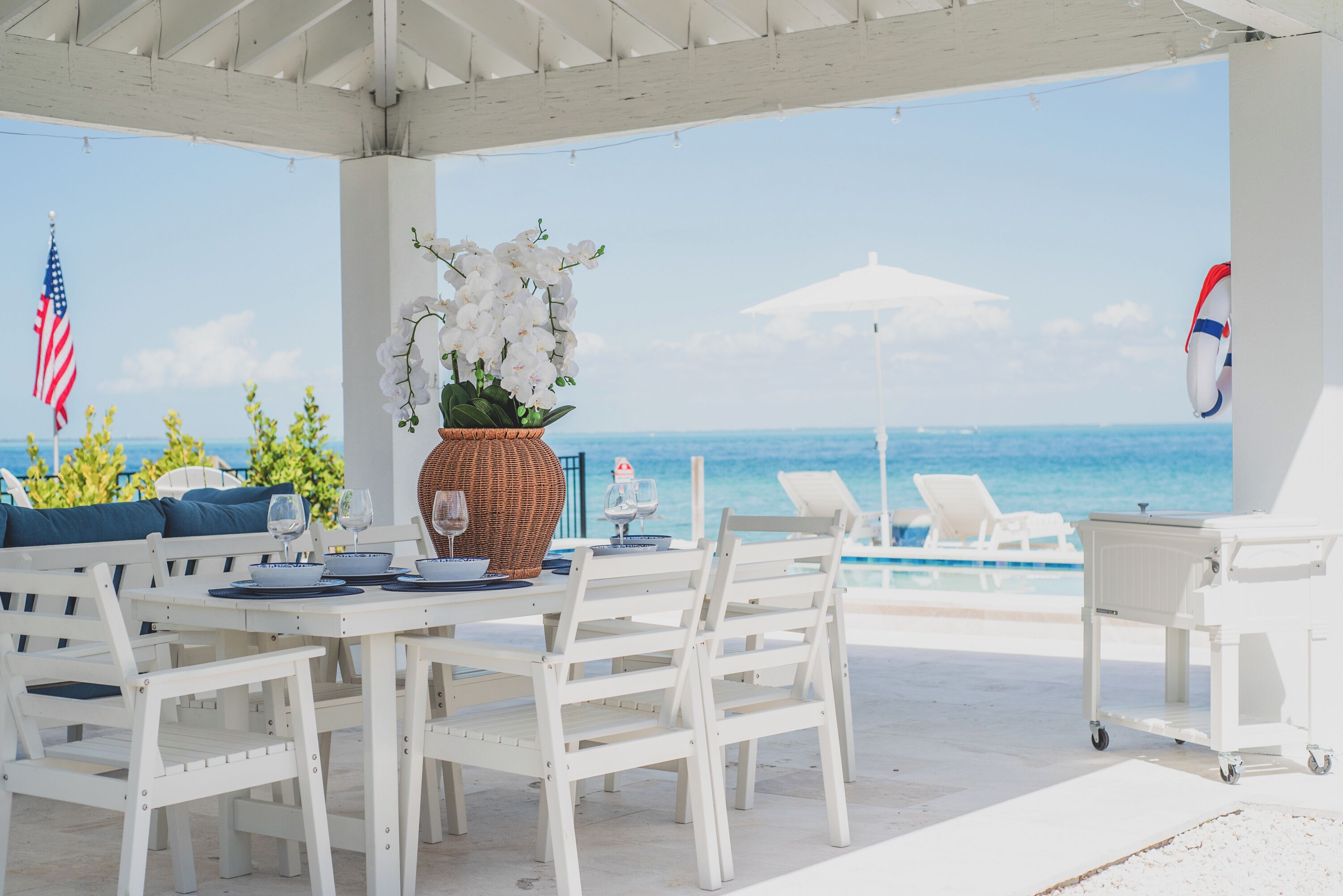 Un área de comedor al aire libre con techo, mobiliario blanco, una mesa con platos y vasos, un centro de mesa de mimbre y vistas al océano.