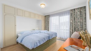 Ein Schlafzimmer mit Bett, einem Holztisch, einer Vase mit Trockenpflanzen und einem Fenster mit Vorhängen.