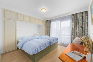 Ein Schlafzimmer mit Bett, einem Holztisch, einer Vase mit Trockenpflanzen und einem Fenster mit Vorhängen.