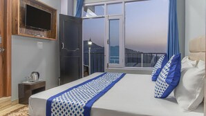 Ένα δωμάτιο ξενοδοχείου με ένα κρεβάτι, μια τηλεόραση τοποθετημένη σε έναν ξύλινο τοίχο, ένα παράθυρο με κουρτίνες και θέα στην πόλη.