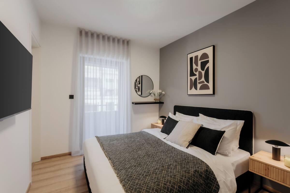A modern bedroom with a large bed, a bedside table with a lamp, a mirror, and a wall art piece.
