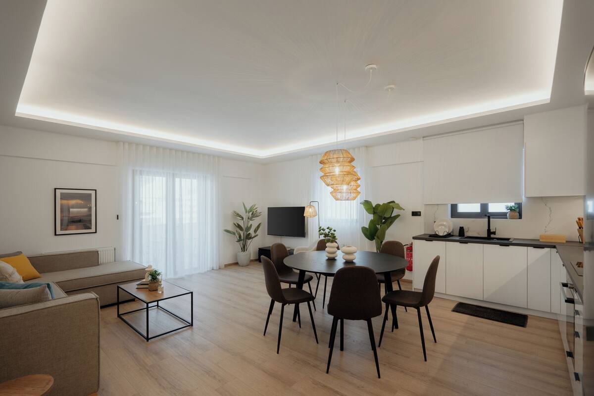 A modern living room with a sofa, coffee table, dining table with chairs, a kitchen area with cabinets, a TV, and a hanging light fixture.