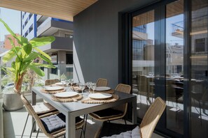 A dining area with a table set for four, surrounded by wicker chairs, a large potted plant, and a view of the city through large windows.