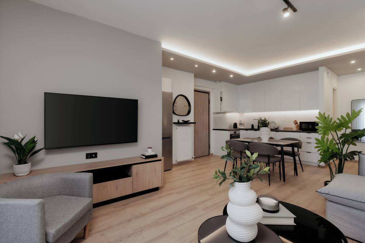 A modern living room with a flat-screen TV, a grey sofa, a dining area with a table and chairs, and a kitchen in the background.