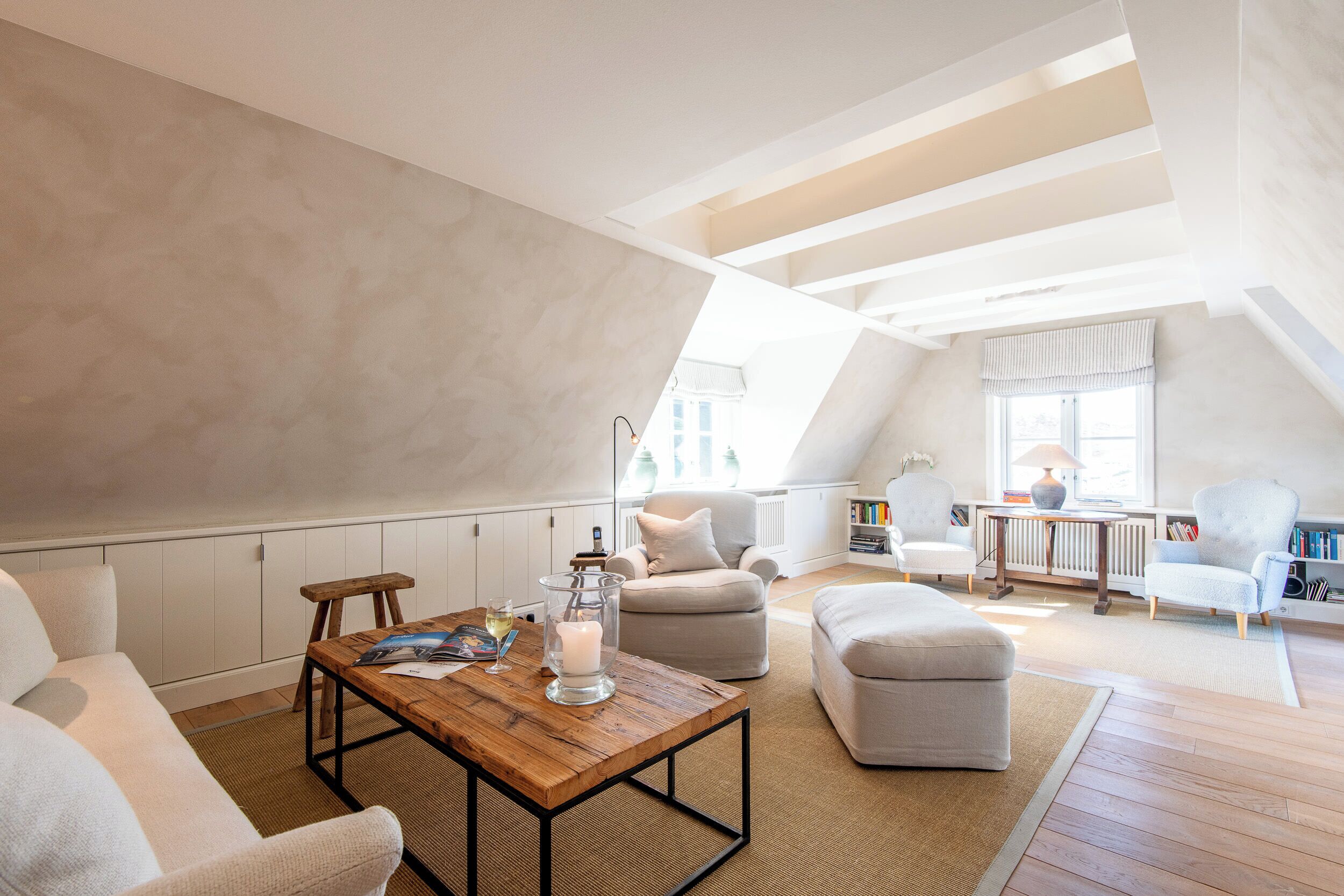Un dormitorio estilo loft con techo alto, vigas a la vista, una mesa de centro de madera, un sofá, un reposapiés y una ventana con vistas.