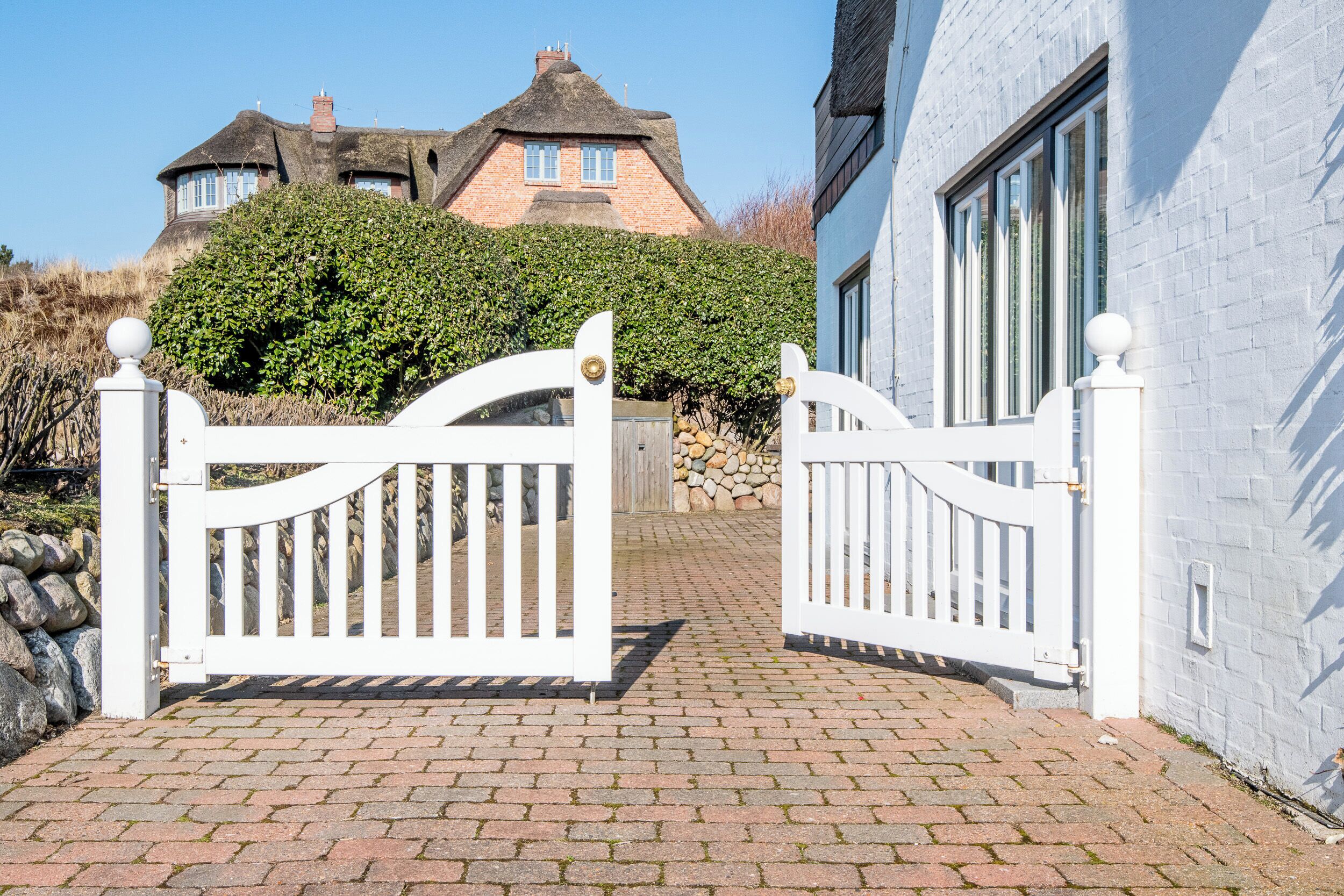 A gated entrance with a brick pathway leading to a house with a thatched roof and a white wall.