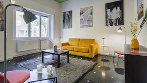 Una sala de estar con un sofá amarillo, una mesa de centro, una mesita con un libro y un vaso, y un jarrón con flores.