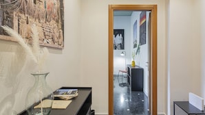 Un pasillo con un espejo, un jarrón con plumas, un escritorio con libros y una puerta que da a otra habitación.