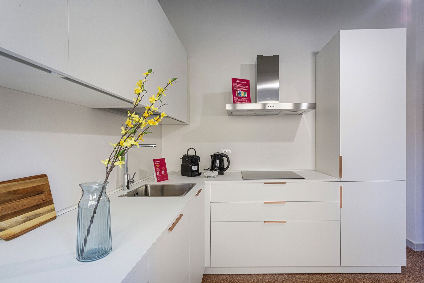 Una cocina moderna con armarios blancos, una campana extractora de acero inoxidable, una cafetera y un florero con flores amarillas.