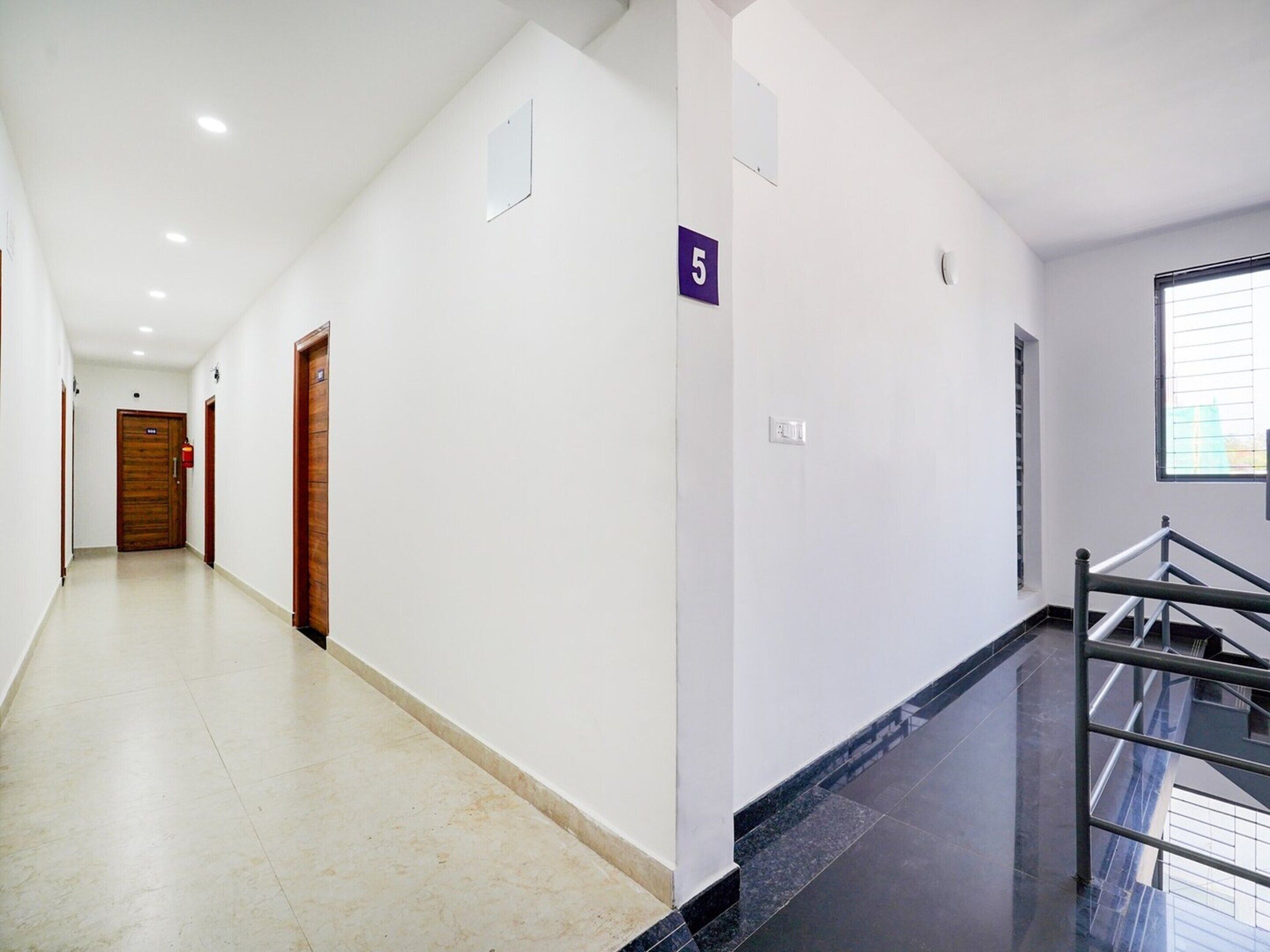A clean, modern hallway with a wooden door at the end, a number '5' sign, and a staircase with metal railings.