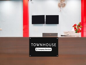A reception desk with a sign reading 'TOWNHOUSE Company-Serviced,' two monitors above it, a fan on the wall, and a vase with flowers on the desk.