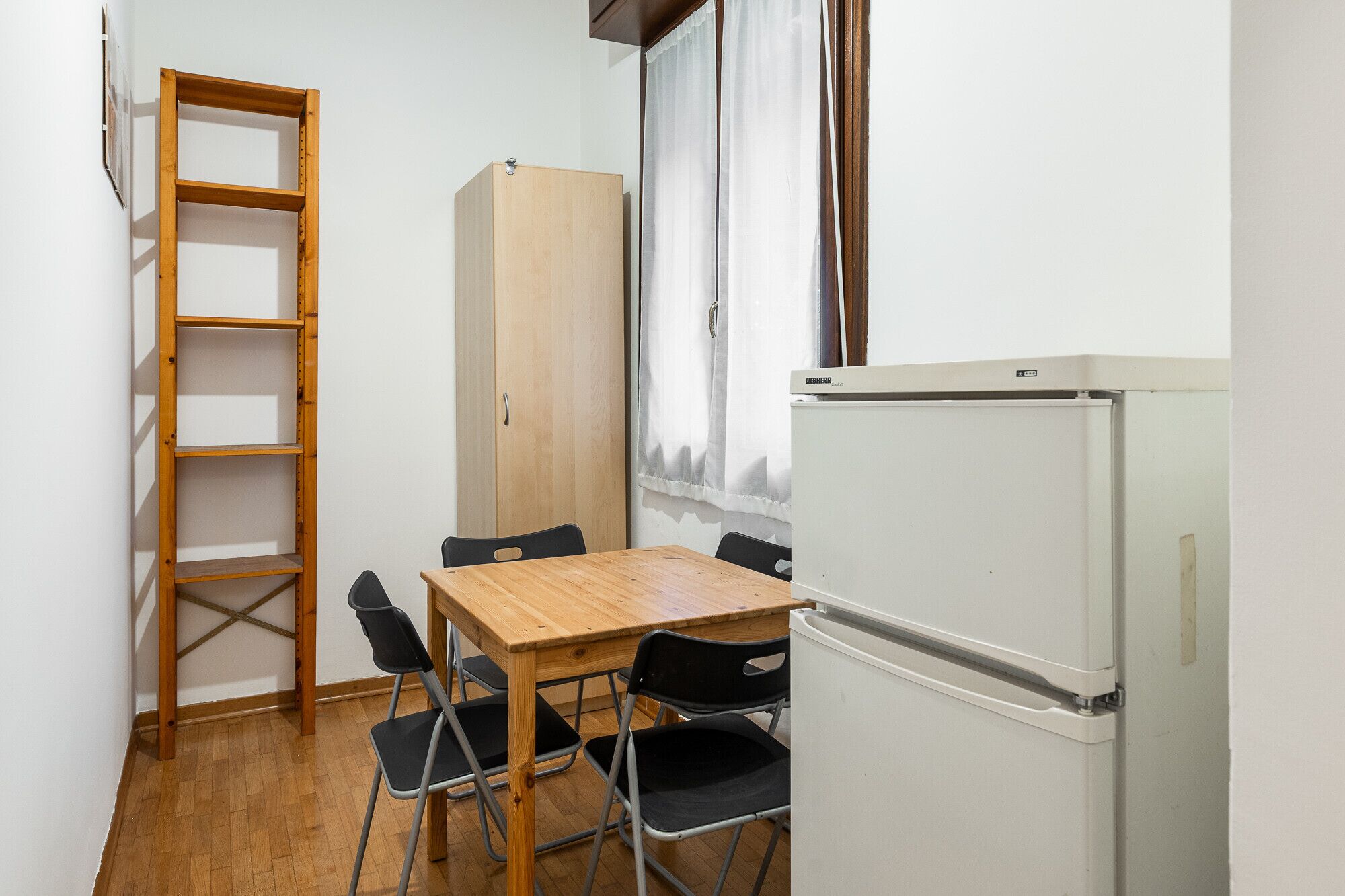 Ein kleines Zimmer mit einem Holztisch, vier Stühlen, einem weißen Kühlschrank, einem Holzschrank und einer Holzleiter an der Wand.