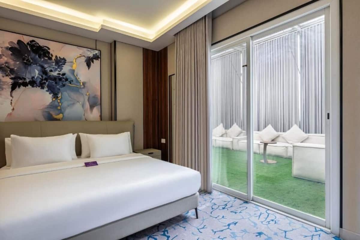 A hotel room with a bed, a large window, a balcony with outdoor seating, and a modern abstract painting on the wall.
