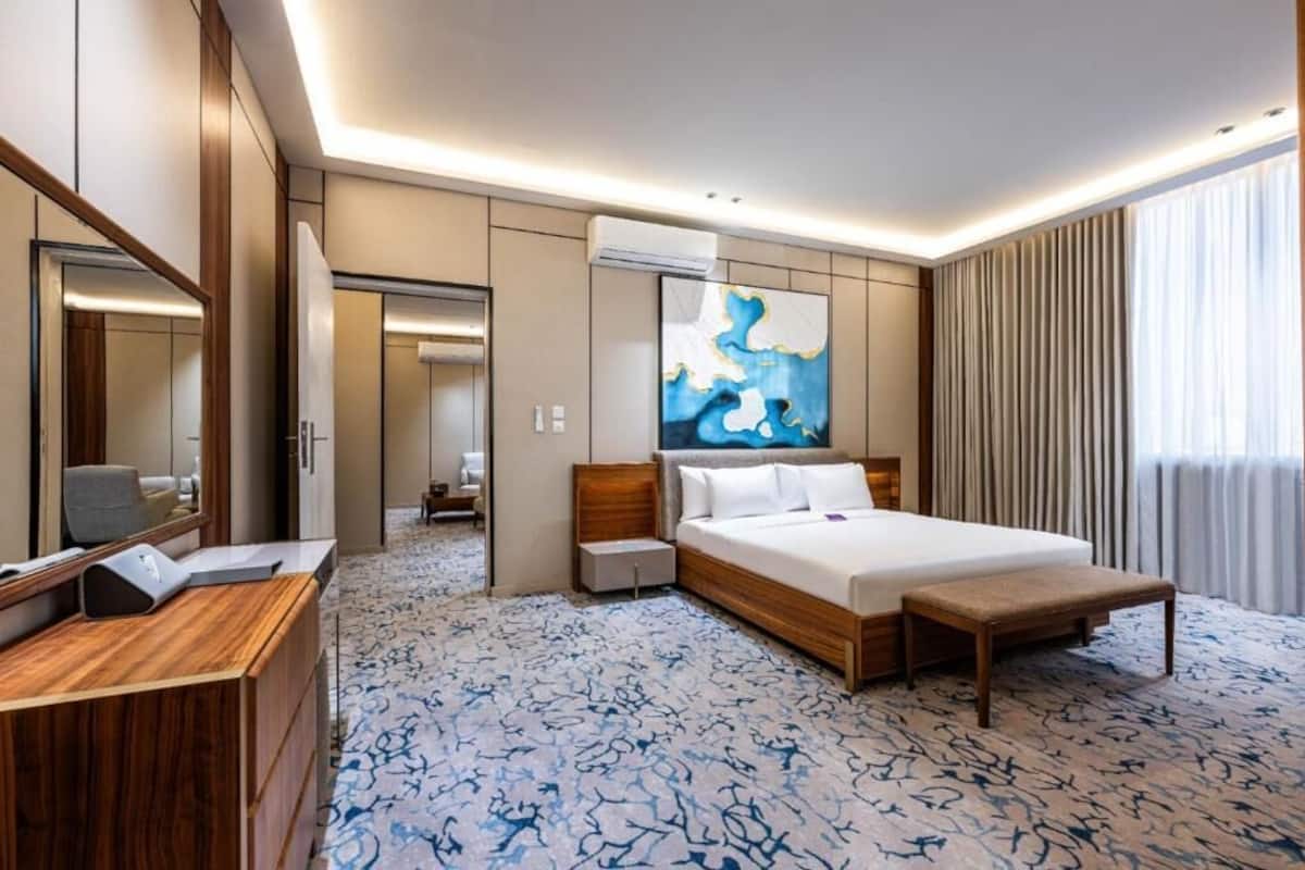 A hotel room with a bed, a desk, a mirror, an air conditioning unit, and a large abstract painting on the wall.